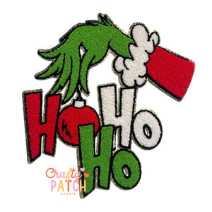 May include: A festive patch featuring a green hand and red sleeve holding a Christmas ornament. The words "Ho Ho Ho" are in red, white, and green, with a gold border. The patch has the text "Crafty Patch Studio".
