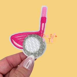 May include: Embroidered patch featuring a pink golf club and a golf ball. The golf club is a vibrant pink with white stitching, and the golf ball is gray with white detailing. The patch is on a yellow background.