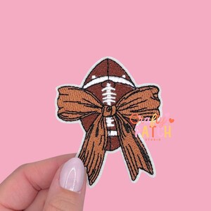 May include: Embroidered patch featuring a brown football with white laces and a large brown bow. The patch has a white border and is set against a pink background. The text "Crafty Patch Studio" is visible.
