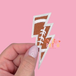 May include: A brown and white embroidered patch in the shape of a lightning bolt, designed to resemble a football. The patch has a white border and features the text "Crafty Patch Studio".