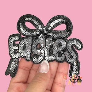 May include: A black sequined patch with the word "EAGLES" in silver sequins, topped with a bow. The patch is on a pink background. The patch is likely for apparel or accessories.
