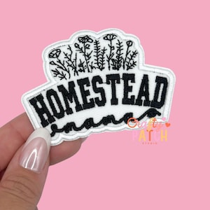 May include: Embroidered patch with the word "HOMESTEAD" in black block letters, outlined in white. The patch features a floral design above the word and a wavy line below. The background is pink.