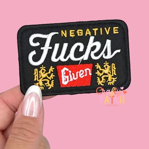 May include: Black rectangular patch with the words "NEGATIVE FUCKS" in white script. The word "Given" is in a red rectangle. Gold lion-like figures flank the sides. The patch is held by a hand.
