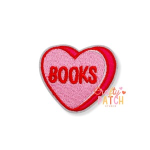 May include: A heart-shaped embroidered patch featuring a light pink center with the word BOOKS stitched in bold red letters. The heart is outlined with a thick red border and a thin grey edge, creating a layered appearance with a vibrant finish.
