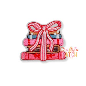 May include: An embroidered patch featuring a stack of four books in shades of tan, blue, pink, and red, tied together with a large, decorative pink and white striped ribbon bow. The patch has a white border and a flat, stitched texture.