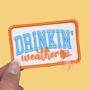 May include: Embroidered patch with the words "DRINKIN' weather" in blue and orange. The patch has an orange border and a small illustration of a drink. The patch is rectangular and is held by a hand.