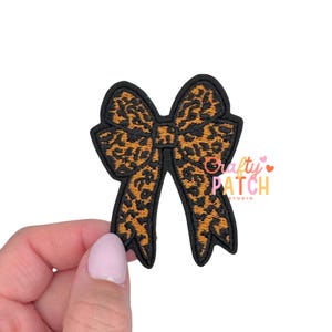 May include: A black-edged bow-shaped patch with a leopard print design in shades of brown and black. The patch is held by a hand against a white background. The text "Crafty Patch Studio" is visible on the right side of the bow.