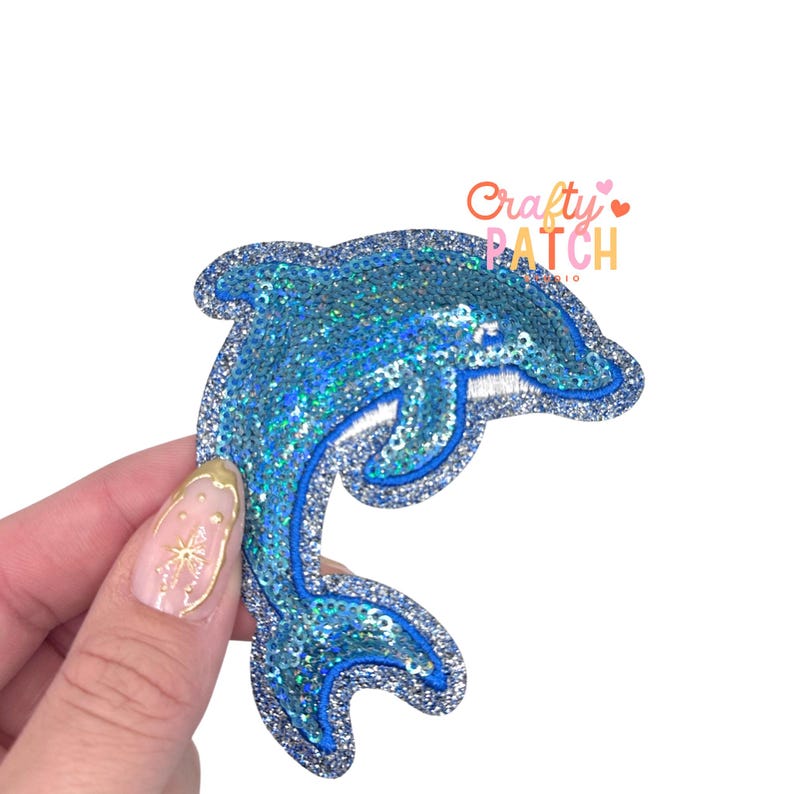 May include: A blue sequined dolphin patch with a silver border. The patch has a glittery, iridescent appearance and is embroidered with white thread. The text "Crafty Patch Studio" is in the upper right corner.