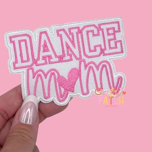 May include: Embroidered patch with the words "DANCE MOM" in pink lettering with a pink heart. The patch has a white border and is on a pink background. The patch is held by a hand.