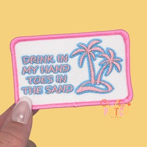 May include: Embroidered patch with a white background and a pink border. The patch features a palm tree design in pink and blue, with the text "DRINK IN MY HAND TOES IN THE SAND" in blue. The patch is held by a hand.