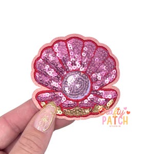 May include: A pink sequined seashell patch with a pearl center and gold accents. The patch has a red outline and the text "Crafty Patch Studio" in pink and orange.