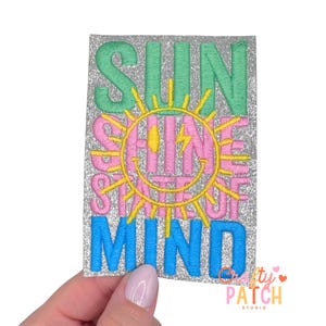 May include: Embroidered patch with the words "SUN SHINE STATE OF MIND" in green, pink, and blue. The design includes a smiling sun with a lightning bolt. The patch has a glittery silver background.