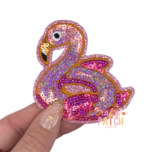 May include: A pink sequined flamingo patch with a gold outline. The patch has a glittery, iridescent effect and features a black and gold beak. The patch is held by a person's hand. The text "Crafty Patch Studio" is visible.