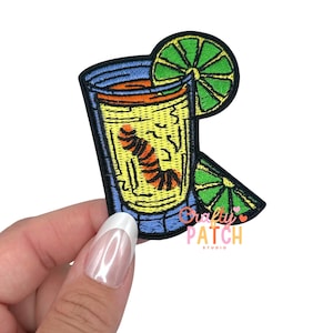 May include: Embroidered patch featuring a cocktail in a glass with a lime wedge. The glass has a yellow drink with an orange worm. The patch has a blue border and the text "Crafty Patch Studio".