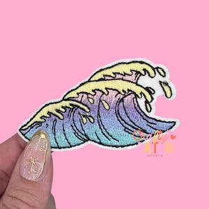 May include: Embroidered patch featuring a stylized ocean wave design. The wave crests are yellow, transitioning to shades of purple and blue. The patch has a white border and is likely for clothing or accessories.