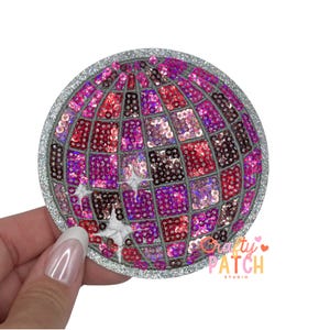May include: A round, sequined patch in the shape of a disco ball. The patch features a silver glitter border and a mosaic of pink, red, and purple sequins. The text "Crafty Patch Studio" is visible on the lower right.