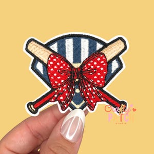 May include: Embroidered patch featuring a red bow with white polka dots, positioned over crossed baseball bats. The bats are tan with red accents. The background is a blue and white striped design, all within a white-bordered shape. The patch has the text "Crafty Patch Studio".