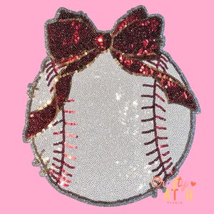 May include: A baseball-shaped decorative item with a large, sequined burgundy bow at the top. The baseball is white with red stitching and a silver sequined border. The background is pink.