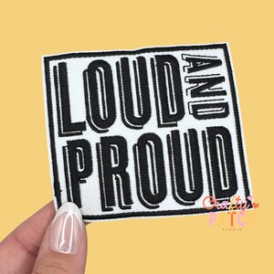 May include: Square white patch with the words "LOUD AND PROUD" in bold, black, outlined letters. The patch is held by a hand with a light yellow background. The patch is a textile item.