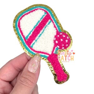 May include: A colorful pickleball paddle patch with a gold glitter border. The paddle features a white center with pink and teal accents. A pink ball is attached to the paddle. The patch is held by a hand.
