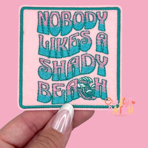 May include: A square, embroidered patch with the text "NOBODY LIKES A SHADY BEACH" in turquoise and pink. The patch has a turquoise border and a pink background. The patch is held by a hand against a pink background.