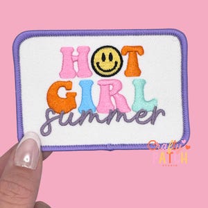 May include: Embroidered patch with the words "HOT GIRL summer" in colorful, bubble letters. The patch features a smiley face and a purple border on a white background. The patch is held by a hand.