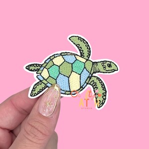 May include: Embroidered sea turtle patch with a geometric design in shades of green, blue, and yellow. The patch has a white border and is set against a pink background. The text "Crafty AT Studio" is visible.