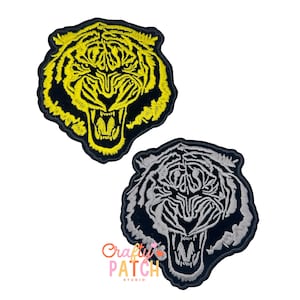 May include: Two embroidered tiger head patches. One patch features a yellow tiger head with black accents, and the other has a gray tiger head with black accents. The patches are on a white background. The patches are from Crafty Patch Studio.