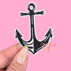 May include: Black embroidered anchor patch with white border, a nautical-themed accessory. The patch features a classic anchor design, perfect for adding to clothing or accessories. The background is a solid pink color.