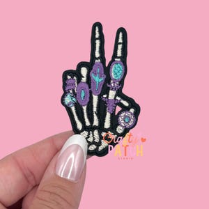 May include: Embroidered patch featuring a black skeleton hand in a peace sign gesture. The fingers are adorned with colorful rings in shades of blue, purple, and teal. The patch has the text "Crafty Patch Studio" in the lower right corner.