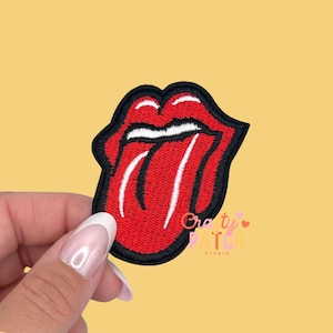 May include: Embroidered patch featuring the iconic red lips and tongue logo with a black border. The patch has white accents and the text "Crafty Patch Studio" in pink and white. The patch is held against a yellow background.