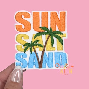 May include: Embroidered patch with the words "SUN SALT SAND" in orange, yellow, and blue. Two palm trees are in the center. The patch is on a white background with a pink background.