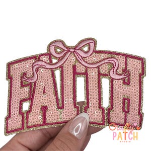 May include: A pink sequined patch with the word "FAITH" in bold, capital letters. The patch has a gold border and a pink bow at the top. The patch is held by a hand.