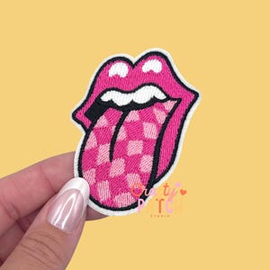 May include: A pink embroidered patch in the shape of a mouth with a tongue sticking out. The tongue has a pink and white checkered pattern. The mouth is outlined in white and black. The patch is held by a hand. The text "Crafty Patch Studio" is in the bottom right corner.
