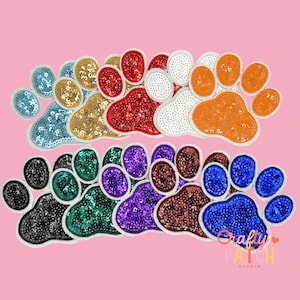 May include: A collection of sequined paw print patches in various colors, including blue, gold, red, white, and orange. The patches are arranged on a pink background. The text "Crafty Patch Studio" is visible in the lower right corner.