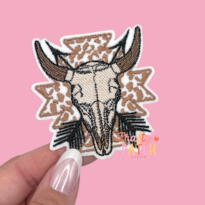 May include: Embroidered patch featuring a bull skull with large horns, set against a geometric pattern with a leopard print design. The patch has a white border and is in shades of brown, black, and beige.