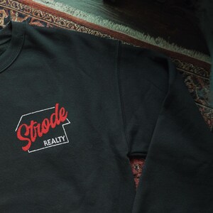 May include: Black crewneck sweatshirt with the red text "Strode Realty" inside a white outlined shape. The sweatshirt is made of a soft material and is suitable for casual wear.