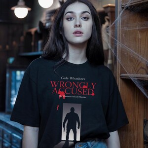 May include: Black t-shirt with the text "Gale Weathers WRONGLY ACCUSED" in red. Below the text is a silhouette of a person holding a knife. The shirt is part of a horror movie-themed collection.