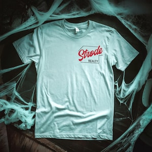 May include: A light blue t-shirt with a red and black logo that says "Strode Realty" in a retro font.