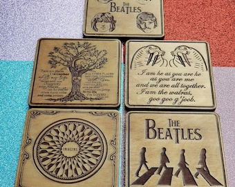 May include: Set of six wooden coasters featuring The Beatles. Each coaster has a different design, including a tree, a walrus, a circle with the word "Imagine", and the Beatles walking across a crosswalk. The coasters are all brown and have a wood grain texture.