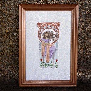May include: A framed embroidery depicting a woman in a purple dress and a peach-colored shawl, sitting on a throne with a circular design behind her. The embroidery is surrounded by a brown frame.