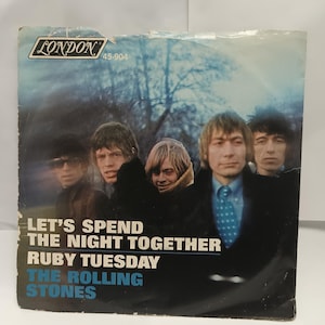 Może przedstawiać: Vintage okładka płyty winylowej z The Rolling Stones. Okładka ma niebiesko-białe tło z twarzami członków zespołu. Tekst na okładce brzmi "Let's Spend The Night Together" i "Ruby Tuesday". Widoczne jest logo London Records.