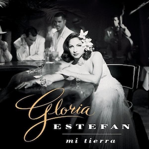May include: Black and white photo of a woman with a flower in her hair, sitting at a bar with a glass of liquid in front of her. The text "Gloria Estefan mi tierra" is written in gold script at the bottom of the image.