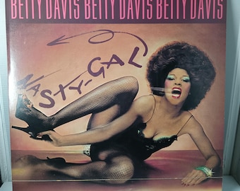May include: Album cover for "Nasty Gal" by Betty Davis. The cover features the artist in a black dress and fishnet stockings, with the album title at the top and the word "Nasty-Gal" in a stylized font.