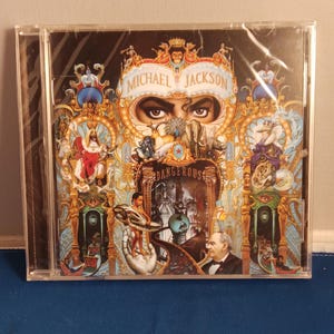 May include: A Michael Jackson Dangerous music CD in a clear plastic jewel case. The cover art features an intricate, colorful illustration with a central pair of eyes, various animals, figures, and ornate architectural details on a black background.