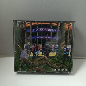 May include: A CD case featuring the Grateful Dead album "Dozin' at the Knick." The cover art depicts a concert scene at Knickerbocker Arena with the band's name prominently displayed. The image has a vintage aesthetic.