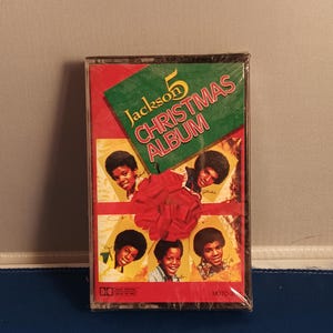 May include: A sealed cassette tape of the Jackson 5 Christmas Album. The cover features a green diagonal banner with the text Jackson 5 CHRISTMAS ALBUM, a large red bow in the center, and four individual portraits of the band members.
