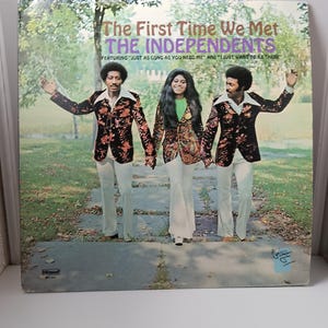 May include: Album cover for "The First Time We Met" by The Independents. The cover features three people holding hands, wearing patterned jackets and white pants. The album title and band name are in purple and pink text.