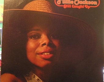 May include: Album cover featuring Millie Jackson, with a large brown hat and a dark background. The text "Millie Jackson Still Caught Up" is in a red and white retro font. The Spring Records logo is in the bottom right corner.