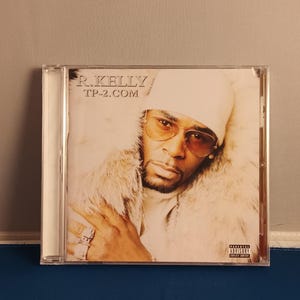 May include: A music CD case featuring the album TP-2.COM by R. Kelly. The cover art displays a portrait of a person wearing a white beanie, tinted glasses, and a white fur-trimmed coat. The text R. KELLY and TP-2.COM appear in the upper left corner.